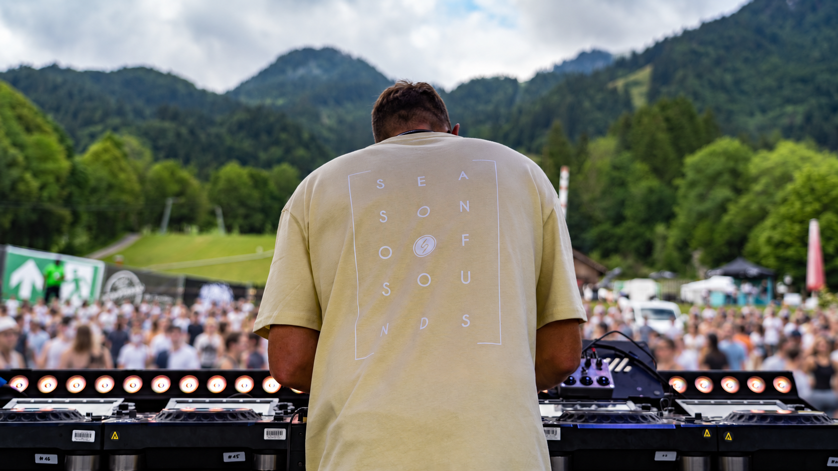 Flow:me hinter dem DJ-Pult mit weich verschwommener Menschenmenge vor dem Bergpanorama